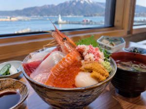 AI-generated illustration: A seafood rice bowl with shrimp, roe and sliced fish beside a harbour-view window.