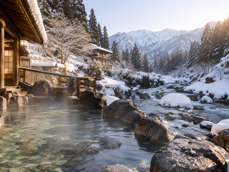 AI-generated illustration: A steaming outdoor hot spring beside a wooden inn, with snow-covered mountains beyond.