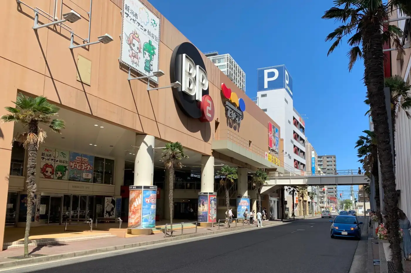 Bandai City BP2 frontage on a sunny day, with the building entrance and palm-lined street.