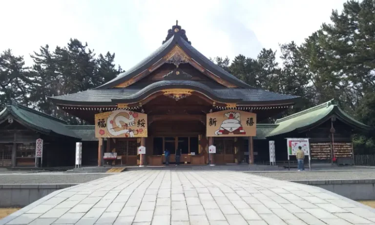 The main shrine hall stands at the end of a broad stone-paved forecourt, with pine trees behind it.