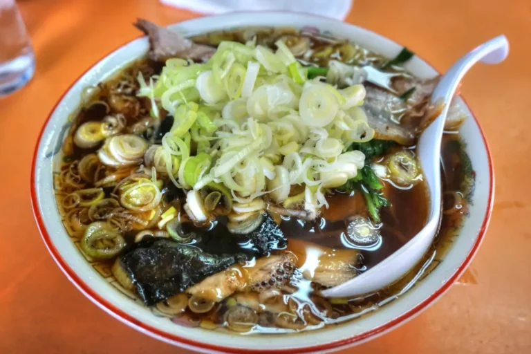 A bowl of dark soy-based ramen topped with sliced pork, chopped scallions, seaweed, and a white soup spoon.