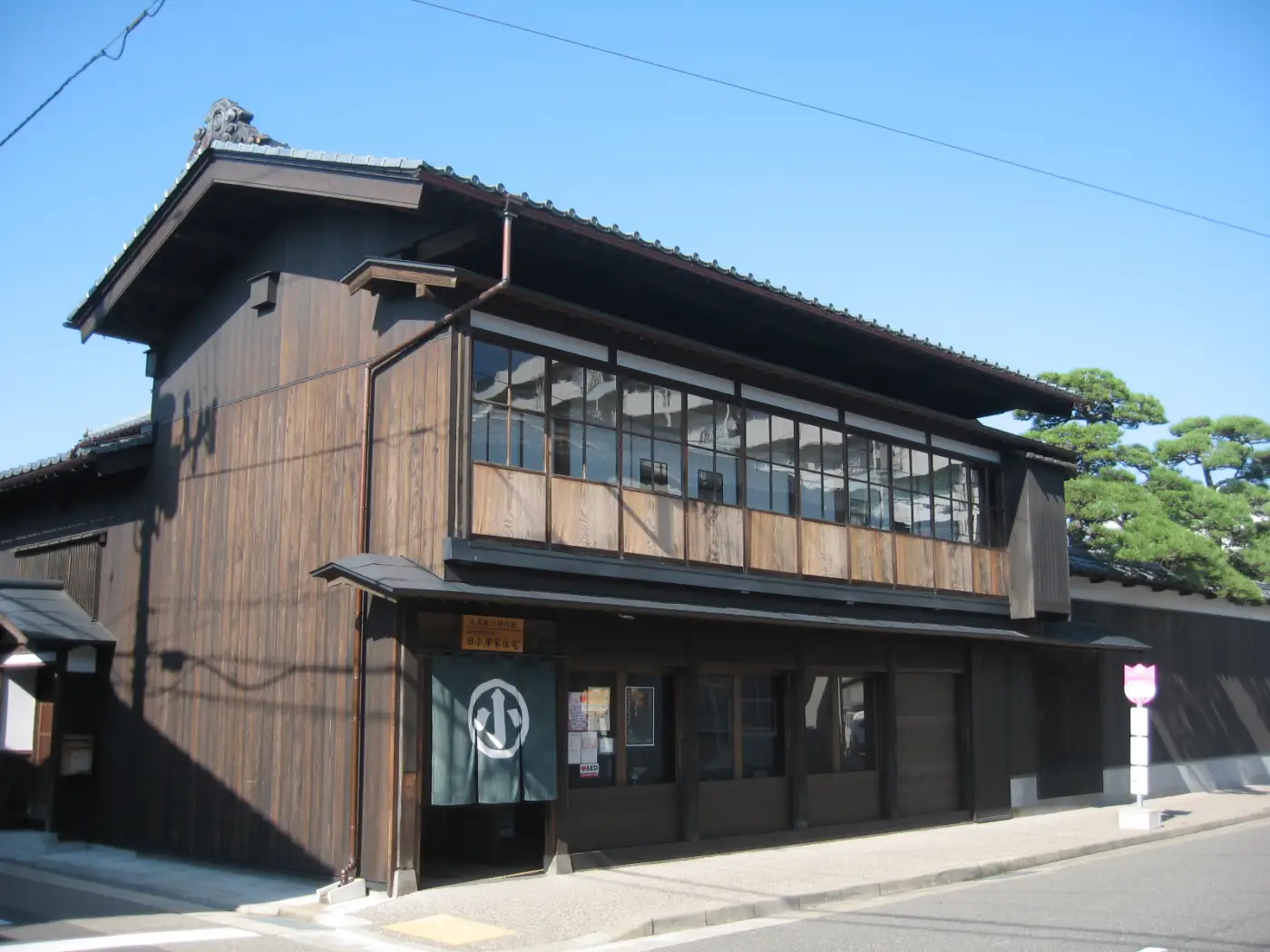The dark wooden façade of the Former Ozawa Residence, with a tiled roof, upper-story windows, and a pine tree behind it.