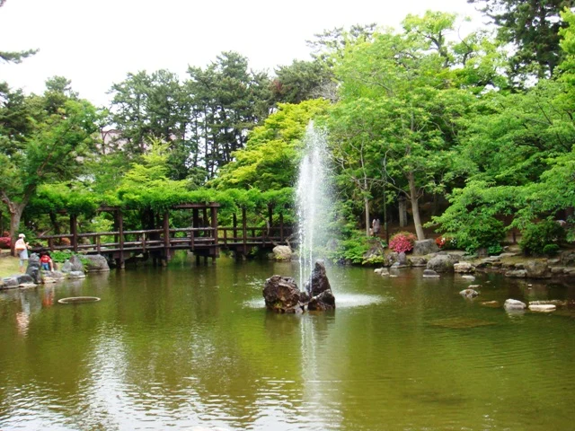 A pond with a central fountain, stepping stones, and a wooden footbridge amid leafy trees.