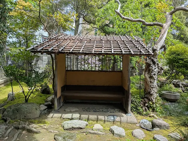A small tiled-roof waiting shelter framed by green garden trees and stones at the Northern Culture Museum Niigata Branch.