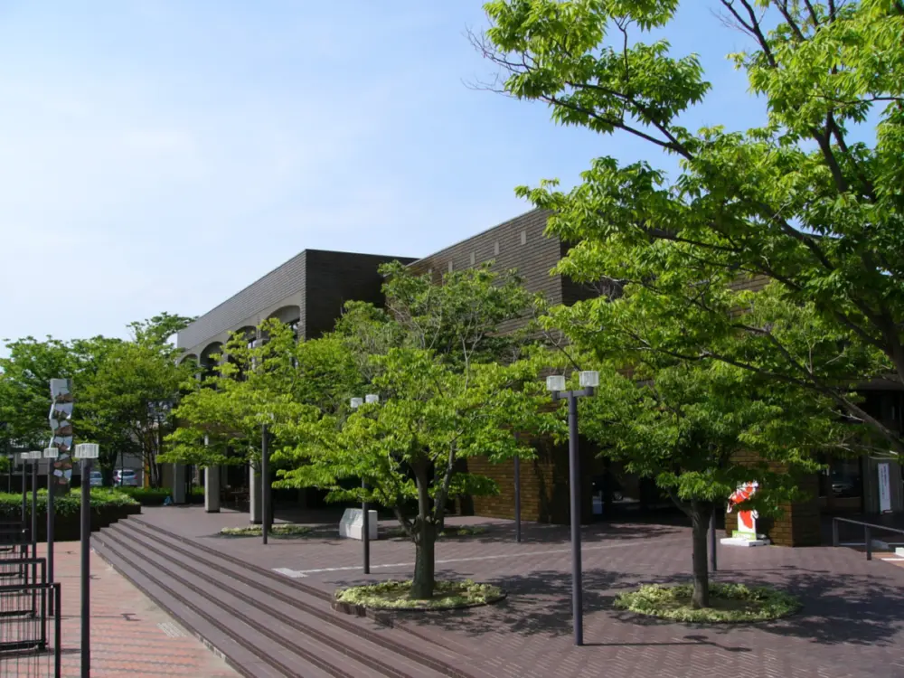 The low, dark-brick Niigata City Art Museum stands behind leafy trees, beside a paved approach and steps.