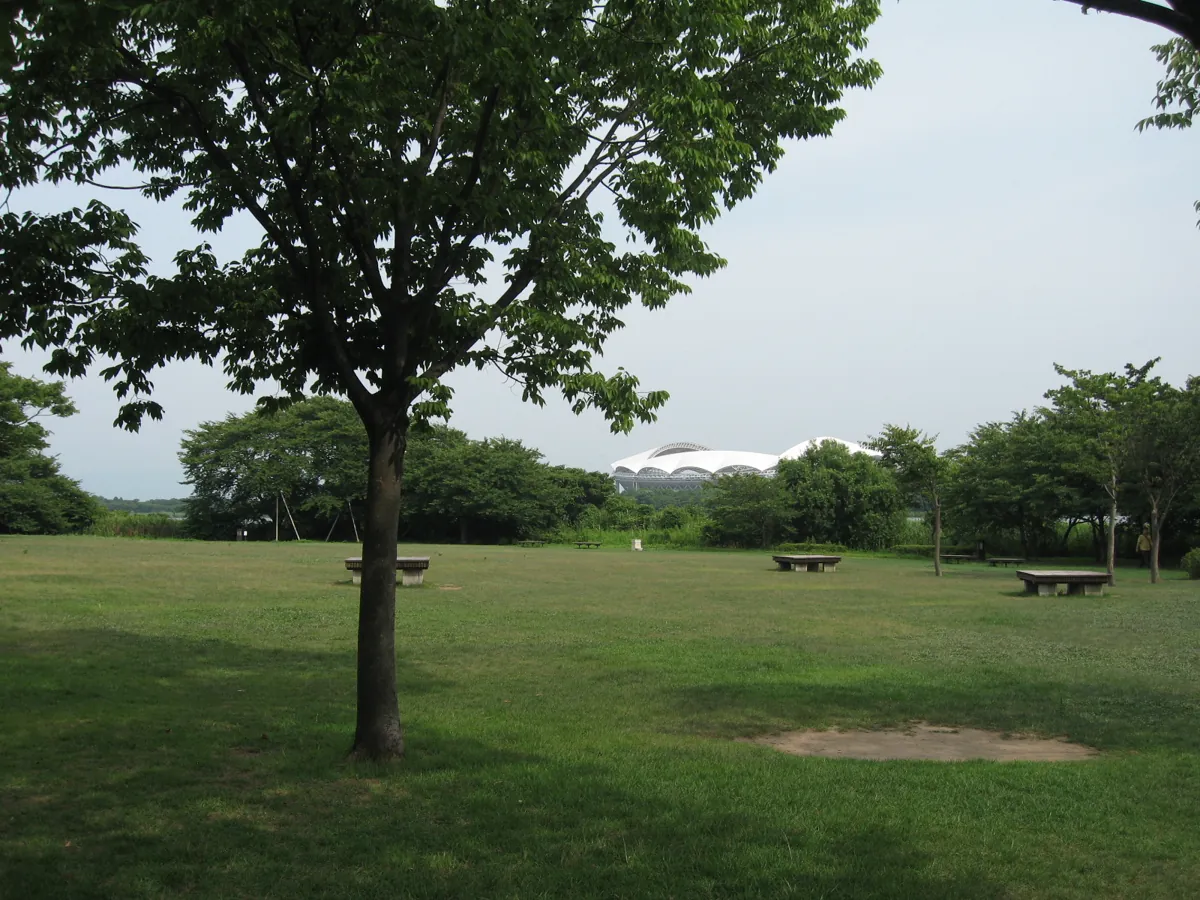Shaded lawn and benches in Toyanogata Park’s Meike district, with Big Swan stadium in the distance.