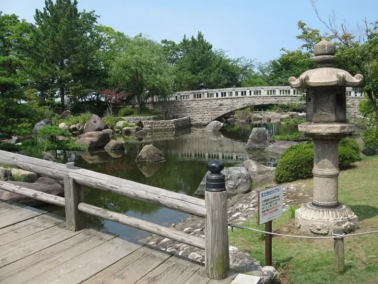 A stone bridge and lantern beside a reflective pond in the Japanese garden of Toyanogata Park’s Shumoku district.